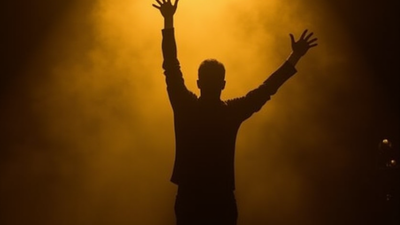 Silhouette of a performer on a dark stage under a golden spotlight with arms raised in triumph
