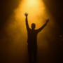 Silhouette of a performer on a dark stage under a golden spotlight with arms raised in triumph