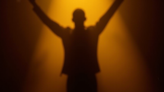 A lone performer silhouetted on a dark stage under a single golden spotlight, arms raised in triumph.