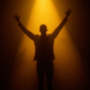 A lone performer silhouetted on a dark stage under a single golden spotlight, arms raised in triumph.