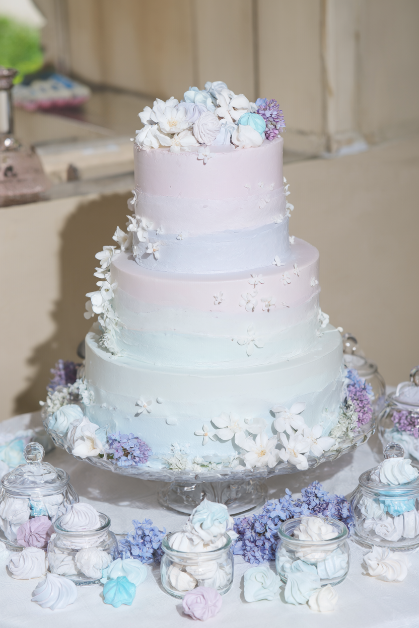 Three-tier cake with lilac to ice-blue ombre buttercream, white sugar blossoms and jars of meringue kisses