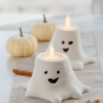Two white air-dry clay ghost tealight holders with painted faces and glowing LED tealights styled on a linen runner with mini pumpkins and autumn leaves