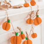 Six small 3D paper pumpkins in warm orange cardstock with brown stems and green leaves strung on a jute twine garland on a white mantel.