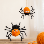 Two plump Halloween pom-pom spiders made of orange and black yarn with pipe-cleaner legs and googly eyes, one on a rustic wood table and one hanging from a cotton string