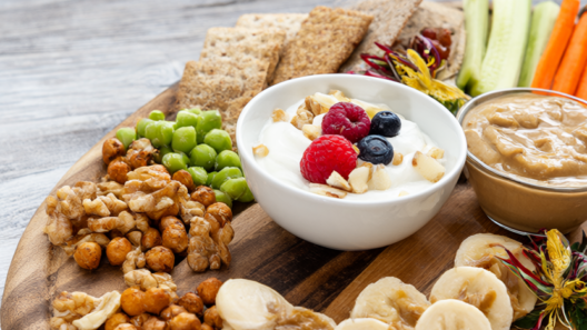 A snack pairing board with Greek yogurt and berries, banana with peanut butter, hummus with veggie sticks, crackers, edamame and roasted chickpeas