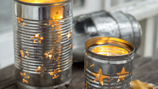 Two upcycled tin can lanterns with punched star and dot patterns glowing from warm LED tea lights on a weathered wooden porch table with dried autumn leaves and twine.