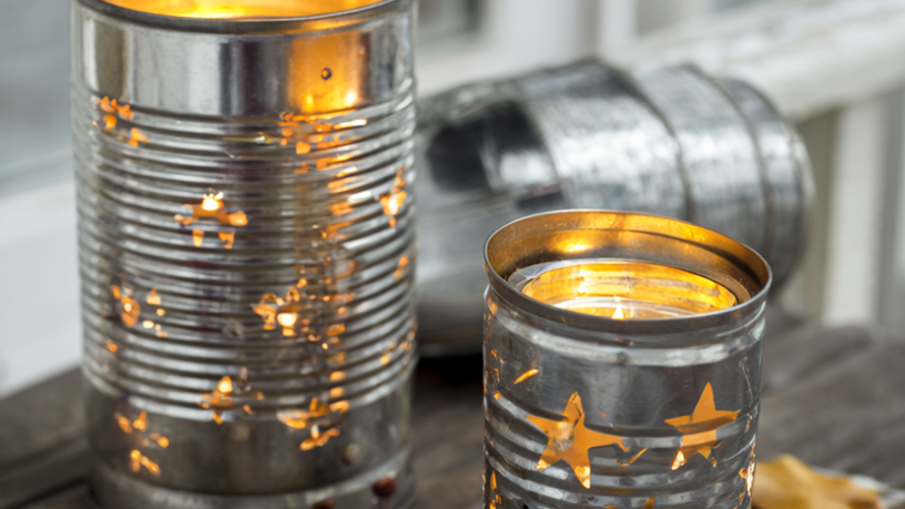 Two upcycled tin can lanterns with punched star and dot patterns glowing from warm LED tea lights on a weathered wooden porch table with dried autumn leaves and twine.
