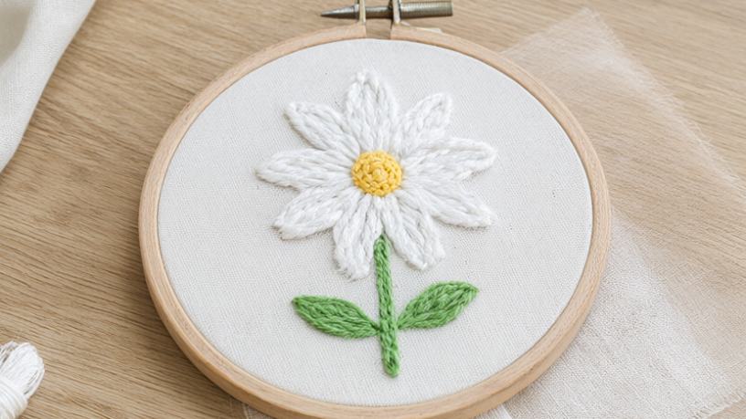 Finished hand-embroidered daisy hoop with white lazy-daisy petals, a butter-yellow French-knot center and green stem and leaves in a 6-inch wooden hoop