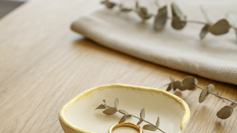 A finished handmade air-dry clay trinket dish in cream clay with a hand-painted gold rim, holding a gold ring and a dried eucalyptus sprig on a light oak table.