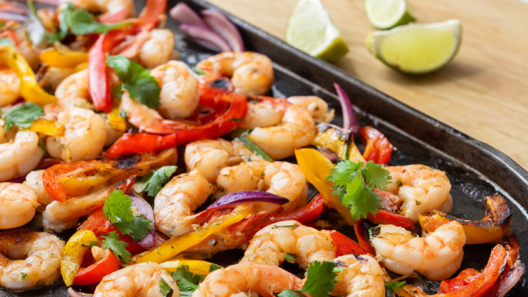Sheet pan shrimp fajitas with charred bell peppers, red onion, cilantro and lime wedges on a dark rimmed baking sheet.