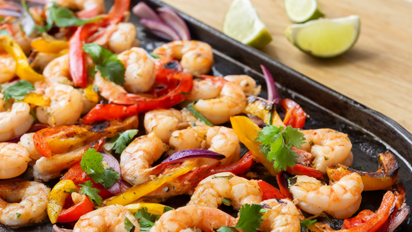 Sheet pan shrimp fajitas with charred bell peppers, red onion, cilantro and lime wedges on a dark rimmed baking sheet.