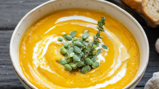 Rustic bowl of silky pumpkin soup with coconut milk, topped with toasted pepitas and a swirl of coconut cream on a dark wooden table.