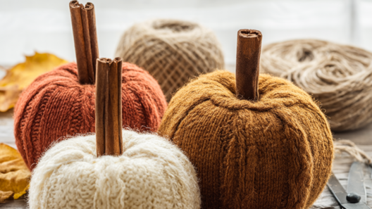 A cluster of three upcycled sweater pumpkins in cream, rust and mustard on a rustic wooden table with cinnamon-stick stems and scattered dried fall leaves