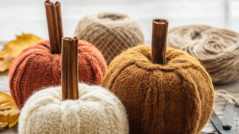 A cluster of three upcycled sweater pumpkins in cream, rust and mustard on a rustic wooden table with cinnamon-stick stems and scattered dried fall leaves