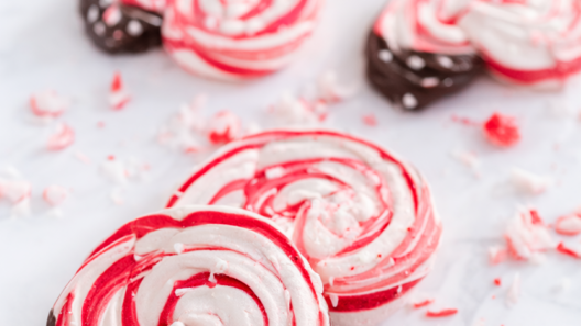Crisp peppermint meringue cookies with red-and-white candy cane swirls and dark chocolate dipped bases on a white marble surface