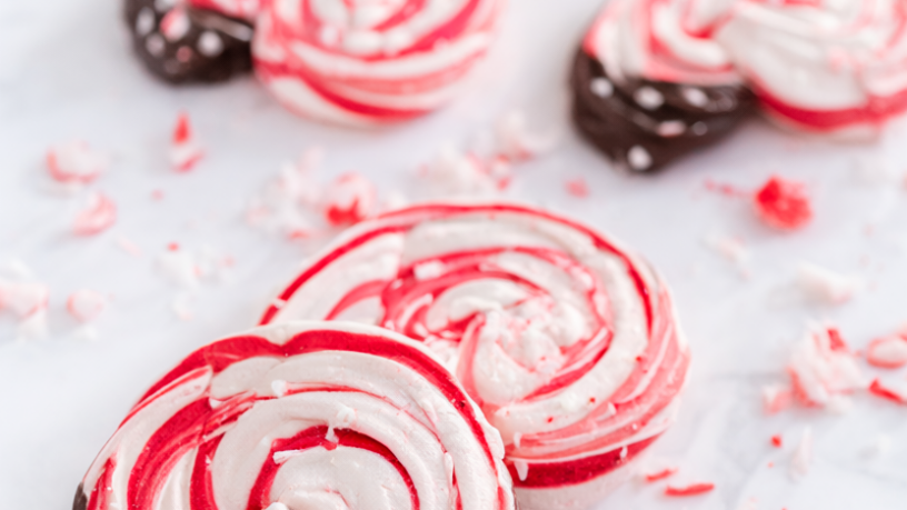 Crisp peppermint meringue cookies with red-and-white candy cane swirls and dark chocolate dipped bases on a white marble surface