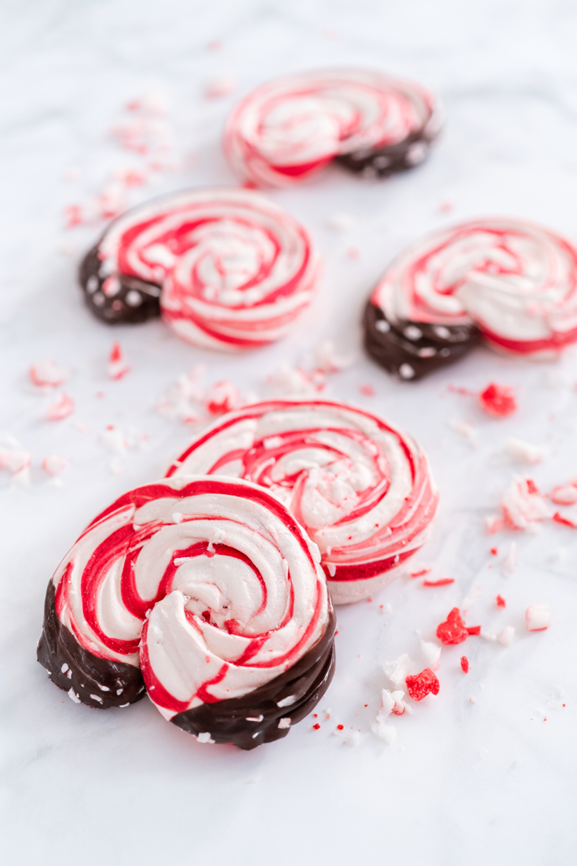Crisp peppermint meringue cookies with red-and-white candy cane swirls and dark chocolate dipped bases on a white marble surface