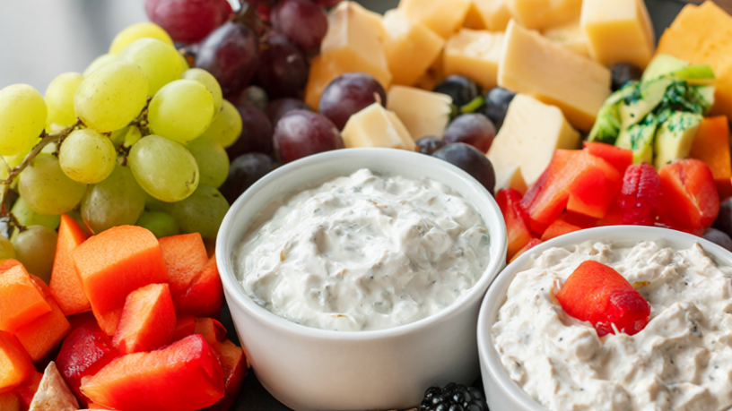 Healthy party snack board with raw vegetables, grapes, berries, cheese, almonds, whole-grain crackers and two bowls of dip.