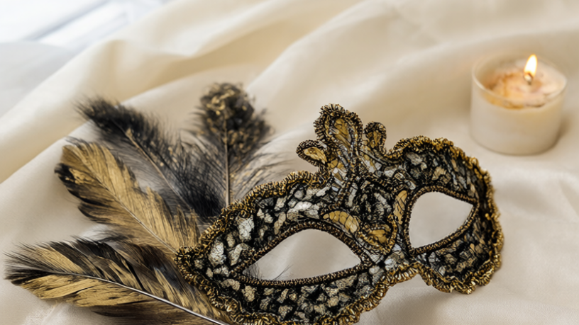 Finished black-and-gold lace masquerade mask with a feather fan and sequins propped on ivory silk by candlelight