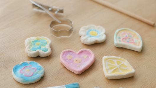 Finished pastel air-dry clay fridge magnets in flower, heart and citrus-slice shapes with painted details on a light oak craft table