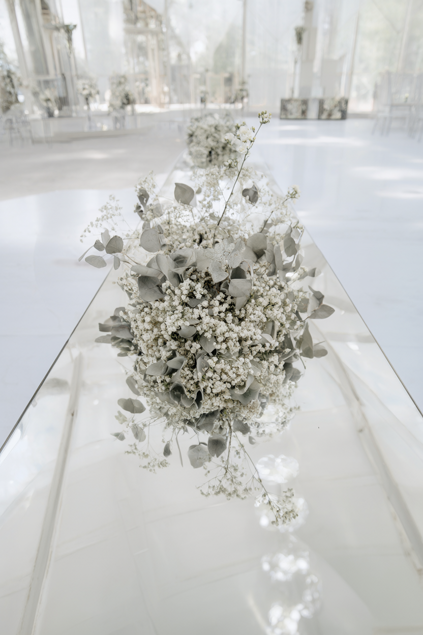 A mirror runner down a white table reflecting small white flower clusters and silver-grey leaves in a bright hall