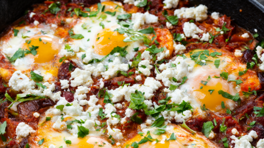 Shakshuka in a black cast-iron skillet: six eggs poached in spiced fresh-tomato pepper sauce, topped with crumbled feta and chopped parsley.