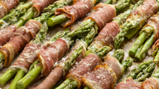Prosciutto wrapped asparagus bundles on a parchment-lined baking sheet with Parmesan, black pepper and lemon wedges.