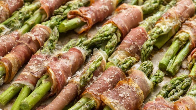 Prosciutto wrapped asparagus bundles on a parchment-lined baking sheet with Parmesan, black pepper and lemon wedges.