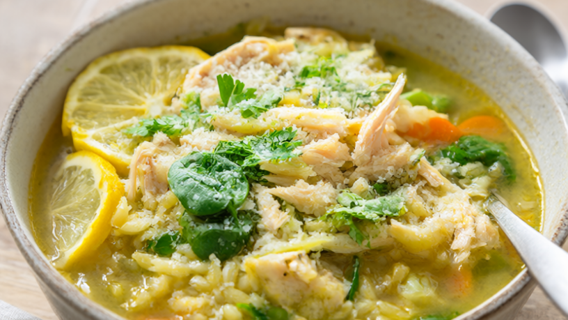 Overhead view of crockpot lemon chicken orzo soup with shredded chicken, spinach, carrots, celery and orzo in a bright lemony broth topped with Parmesan and lemon slices.