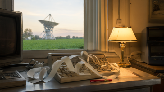 A 1970s radio observatory control room at dawn, with continuous-feed computer paper curling across a metal desk beside a red pen and a tall radio telescope in a green Ohio field outside the window.
