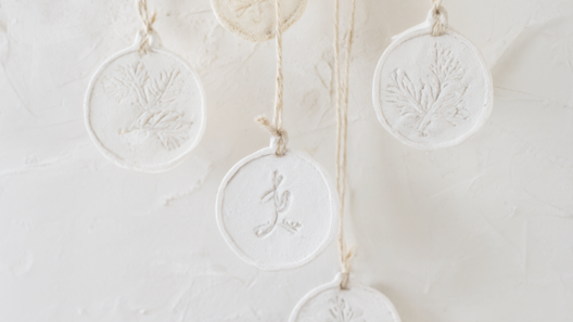 Five finished white air-dry clay Christmas ornaments stamped with pine sprigs and initials, threaded on cream twine against a warm white plaster wall.