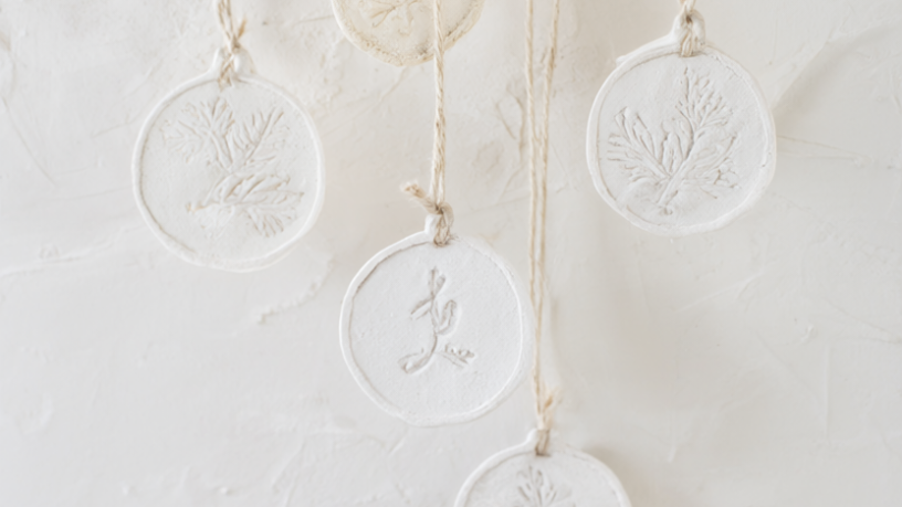 Five finished white air-dry clay Christmas ornaments stamped with pine sprigs and initials, threaded on cream twine against a warm white plaster wall.