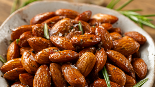 Golden roasted almonds with rosemary, garlic, and sea salt in a rustic ceramic bowl on a wooden table.