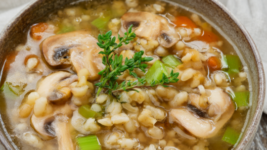 Rustic bowl of mushroom barley soup with cremini mushrooms, pearl barley, carrots, celery, and fresh thyme on a wooden table.