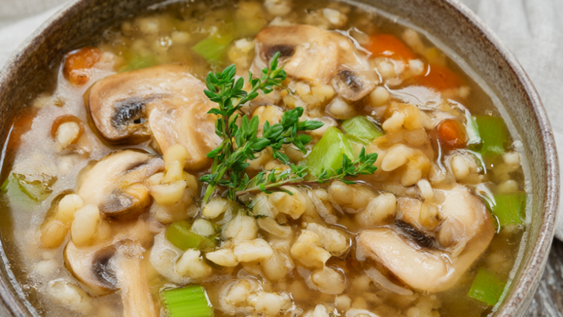 Rustic bowl of mushroom barley soup with cremini mushrooms, pearl barley, carrots, celery, and fresh thyme on a wooden table.