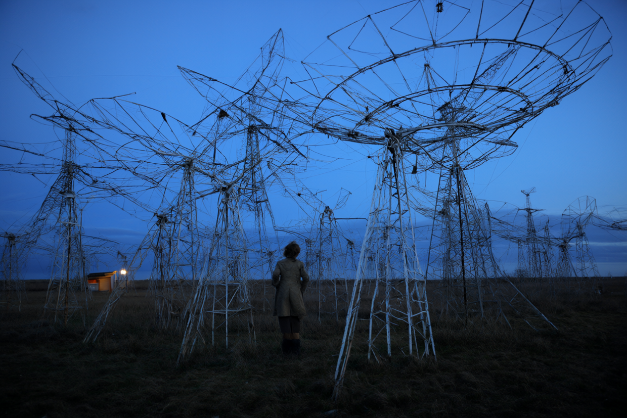 A young woman in a coat stands among wire-strung radio telescope antennae on an English field under a deep blue evening sky.