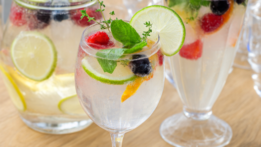 Two sparkling-water mocktails with lime, mint, thyme and frozen berries beside a club soda pitcher on light oak
