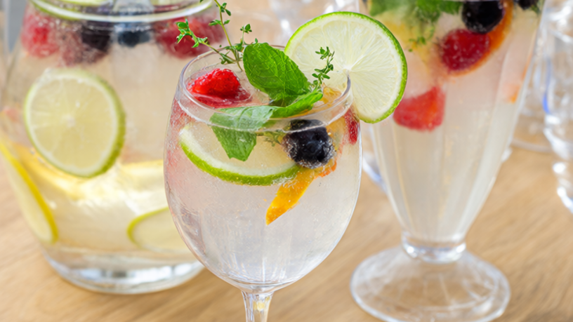 Two sparkling-water mocktails with lime, mint, thyme and frozen berries beside a club soda pitcher on light oak