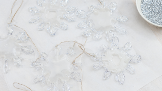 Six translucent hot glue snowflake ornaments edged with silver glitter and hung from twine on a white oak table