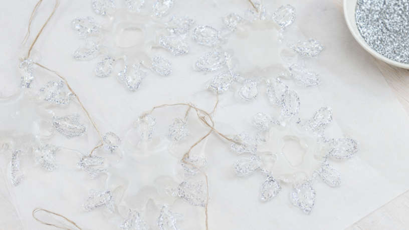 Six translucent hot glue snowflake ornaments edged with silver glitter and hung from twine on a white oak table