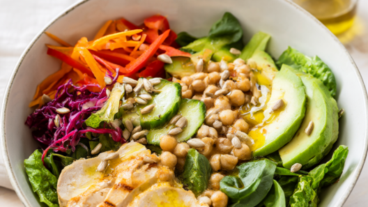Lunch salad bowl with romaine, spinach, grilled chicken, chickpeas, cucumber, carrot, red pepper, cabbage, avocado, sunflower seeds, and vinaigrette.