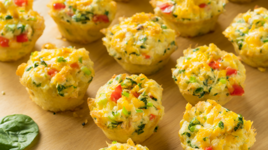 Golden baked egg muffins with spinach, red bell pepper and melted cheddar on a light wood board.