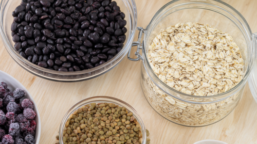 Overhead spread of high-fiber staples: black beans, lentils, rolled oats, almonds, chia seeds, frozen berries, air-popped popcorn and fresh spinach on light wood.