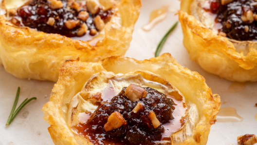 Golden puff pastry brie bites with fig jam, chopped pecans, rosemary and a honey drizzle on a white marble board.