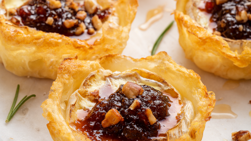 Golden puff pastry brie bites with fig jam, chopped pecans, rosemary and a honey drizzle on a white marble board.