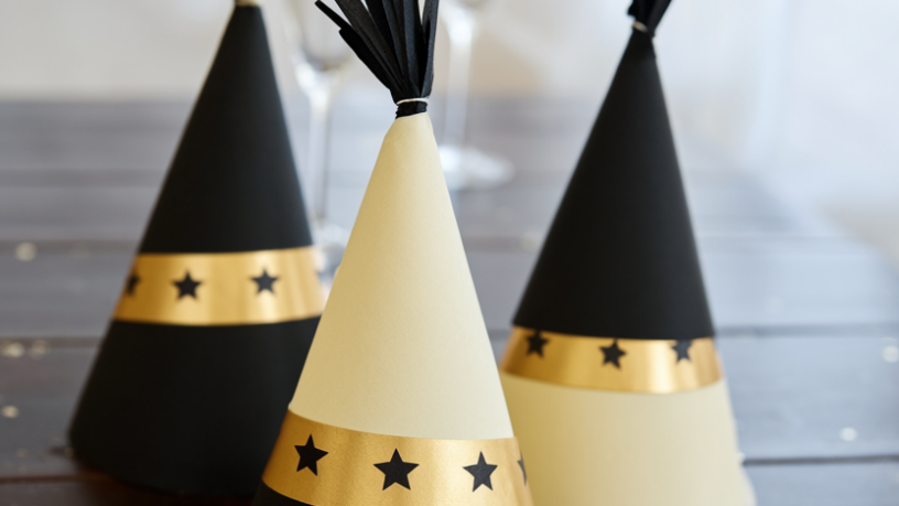 Three tall paper cone party hats in black, cream and gold with star bands and tissue tassel tips on a dark wood table, gold confetti and champagne flutes softly blurred behind them in warm evening light
