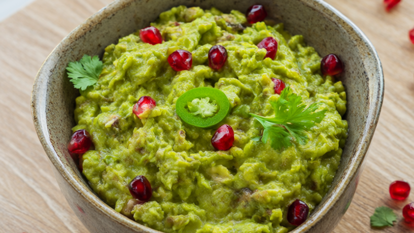 Chunky guacamole topped with pomegranate arils, cilantro and a lime wedge in a ceramic serving bowl