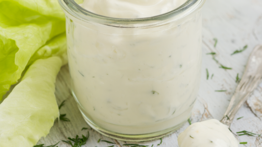 Creamy Greek yogurt dressing in a glass jar with a drizzle spoon, chopped dill and parsley, and pale butter lettuce on rustic wood