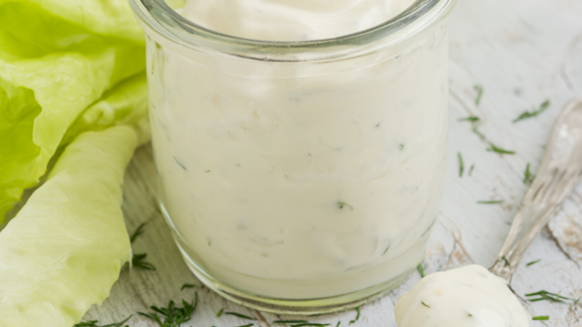 Creamy Greek yogurt dressing in a glass jar with a drizzle spoon, chopped dill and parsley, and pale butter lettuce on rustic wood