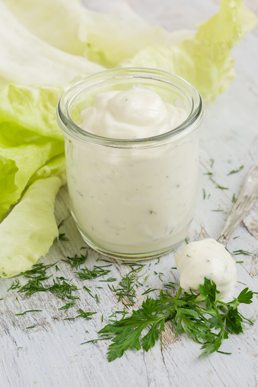 Creamy Greek yogurt dressing in a glass jar with a drizzle spoon, chopped dill and parsley, and pale butter lettuce on rustic wood